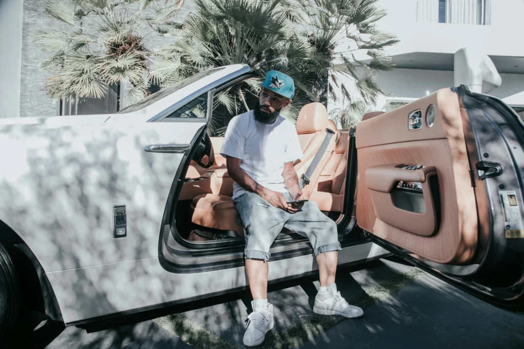 Homem sentado em um Rolls-Royce conversível branco com interior caramelo, capturada sob a sombra de palmeiras. Foto por Unsplash.